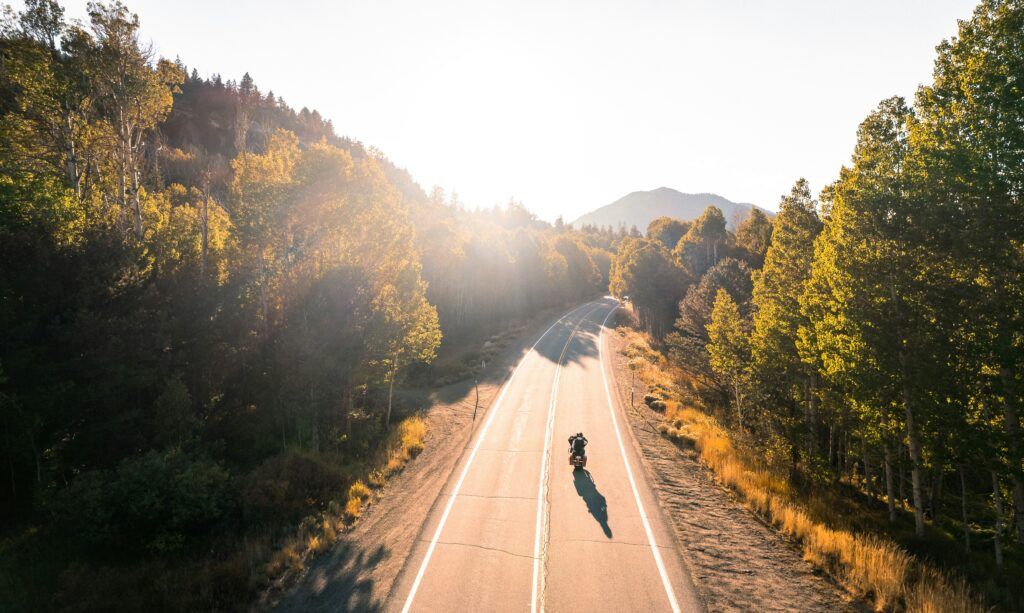 A person riding a motorcycle along a winding mountain road, surrounded by lush greenery and rocky peaks.