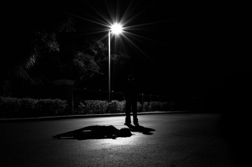 A person standing on a dimly lit street at night, surrounded by shadows and faint streetlights.
