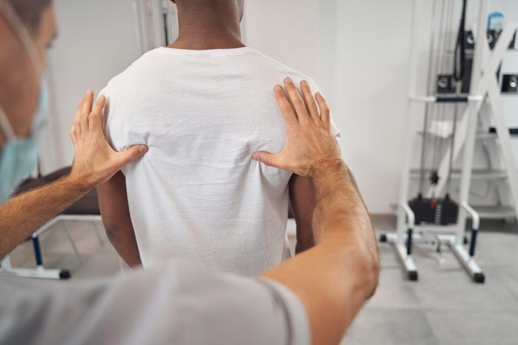 Back view of doctor in face mask palpating male patient paraspinal muscles