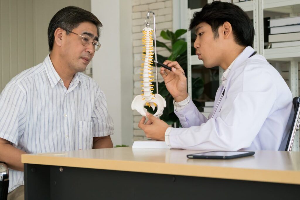 Young male doctor explaining anatomical spine to senior man patient at hospital room.