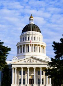 Front view of the California State Capitol building in Sacramento, featuring a prominent white dome with a dark roof, Corinthian columns, neoclassical sculptures, and the American flag at half-staff against a partly cloudy sky.