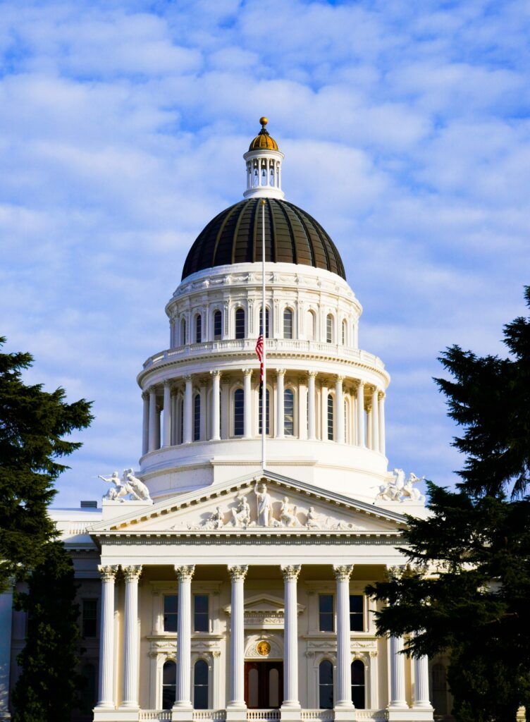 Front view of the California State Capitol building in Sacramento, featuring a prominent white dome with a dark roof, Corinthian columns, neoclassical sculptures, and the American flag at half-staff against a partly cloudy sky.