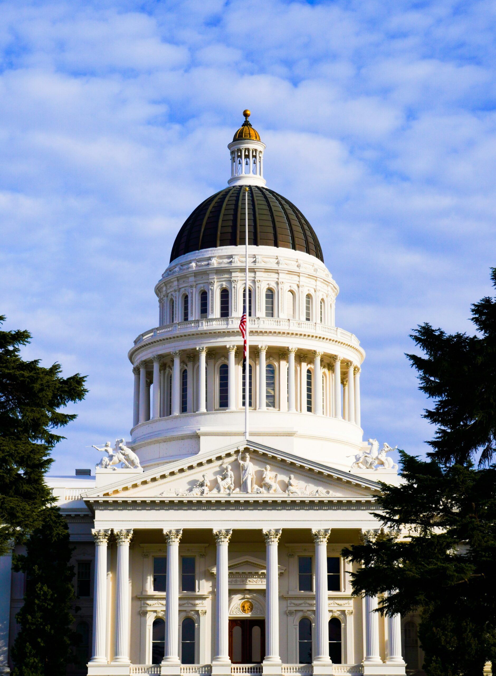 Front view of the California State Capitol building in Sacramento, featuring a prominent white dome with a dark roof, Corinthian columns, neoclassical sculptures, and the American flag at half-staff against a partly cloudy sky.