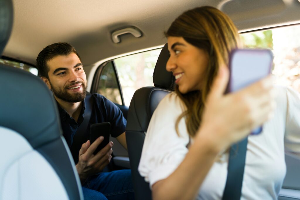 a smiling Uber driver wth a phone in her hand turns to look at a smiling passenger behind her