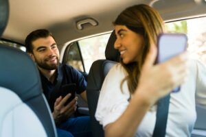 a smiling Uber driver wth a phone in her hand turns to look at a smiling passenger behind her