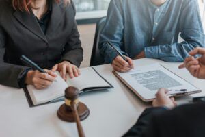 Lawyer meeting with clients at a desk, reviewing legal documents and taking notes. A judge’s gavel rests on the table, symbolizing law and justice during a legal consultation or contract discussion.