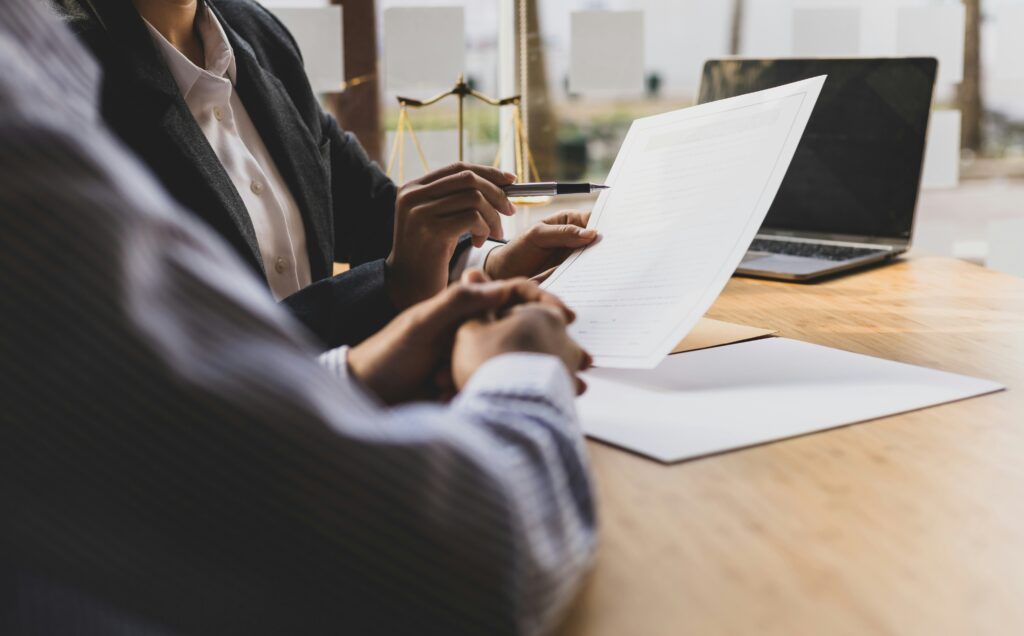Two people in a legal consultation reviewing a document together at a wooden desk, with a laptop and scales of justice in the background, representing professional legal advice and case review.