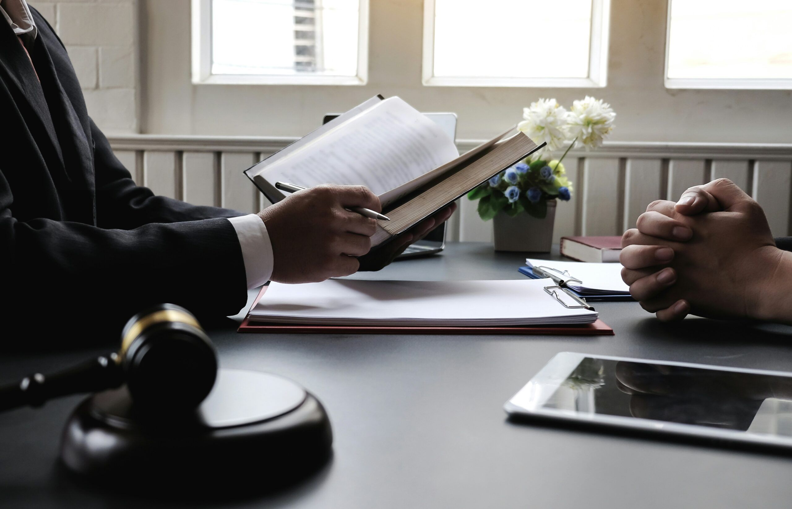 Close-up of a lawyer reading a legal book and consulting with a client across a desk, with documents, a gavel, and a tablet nearby, symbolizing legal guidance and case preparation in a professional office environment.