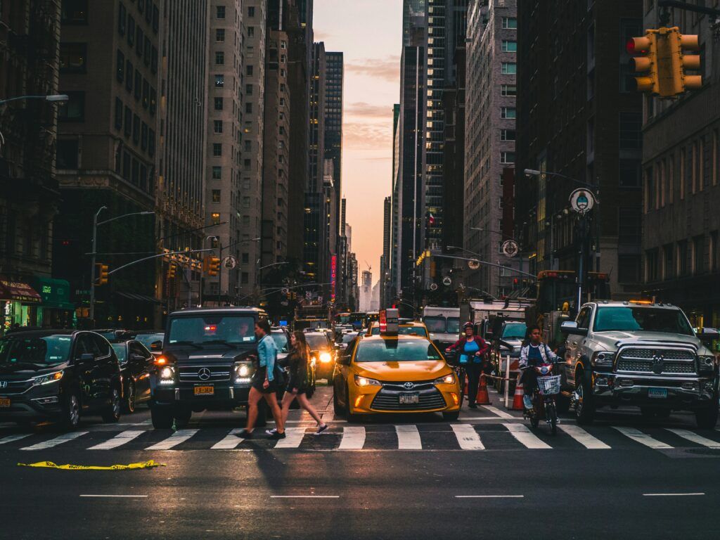 A bustling city intersection at dusk with dense traffic and several pedestrians crossing a wide crosswalk, surrounded by tall buildings and illuminated by streetlights and headlights.