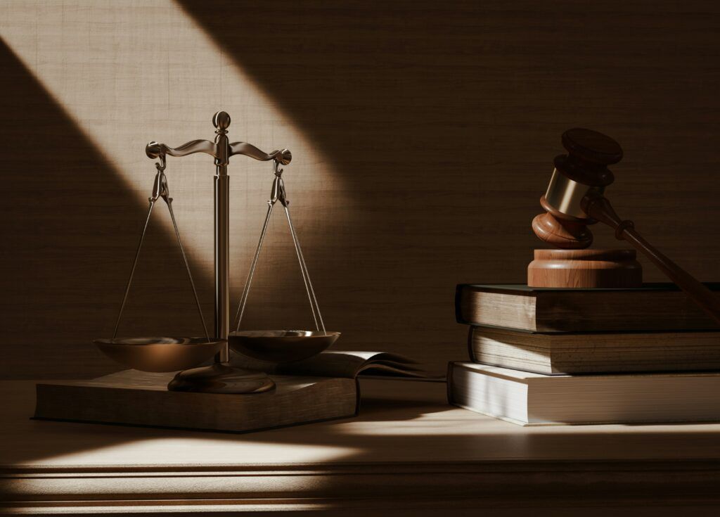 Bronze scales of justice and a wooden gavel resting on top of stacked law books, illuminated by a beam of light in a dimly lit courtroom or legal office, symbolizing fairness and the pursuit of justice.