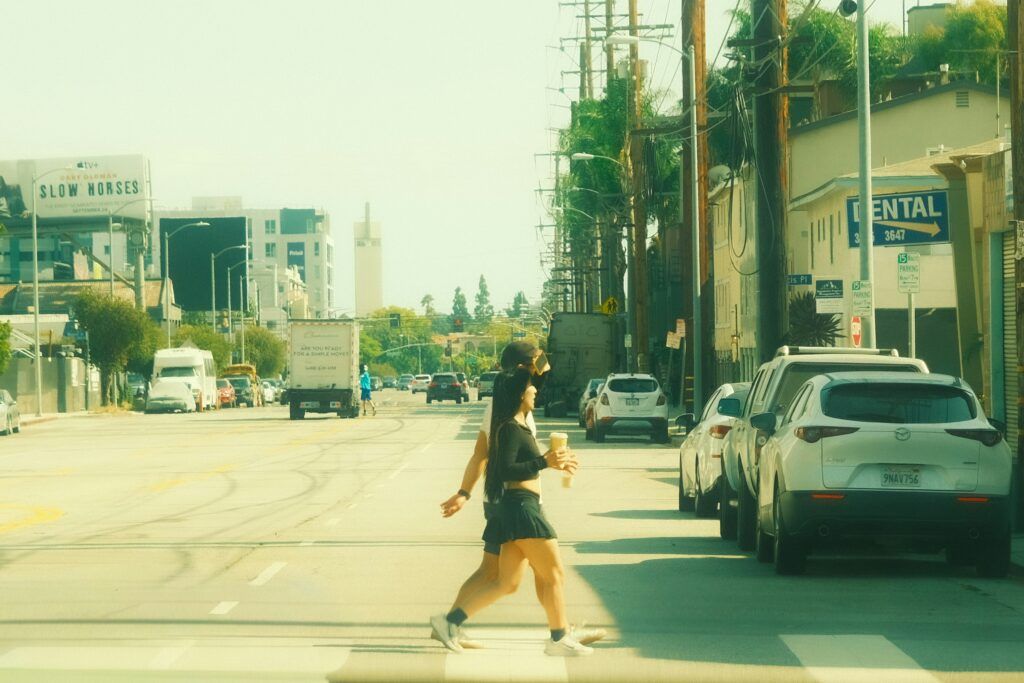 Two pedestrians crossing a sunlit urban street, holding drinks, with parked cars lining the road and city buildings in the background