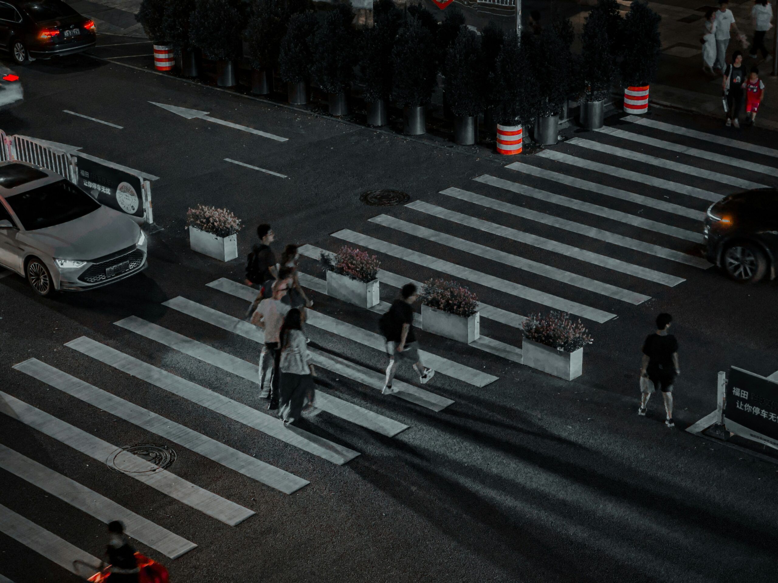 A nighttime urban crosswalk scene with several pedestrians walking across wide, white-striped crosswalks, illuminated by streetlights and passing vehicles, with planters and traffic cones placed between the lanes.