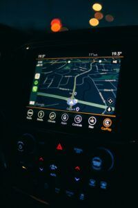 A car’s dashboard navigation screen at night showing a digital map with streets and route guidance, illuminated controls below the display, temperature and direction indicators at the top, and blurred city lights visible through the windshield in the background.