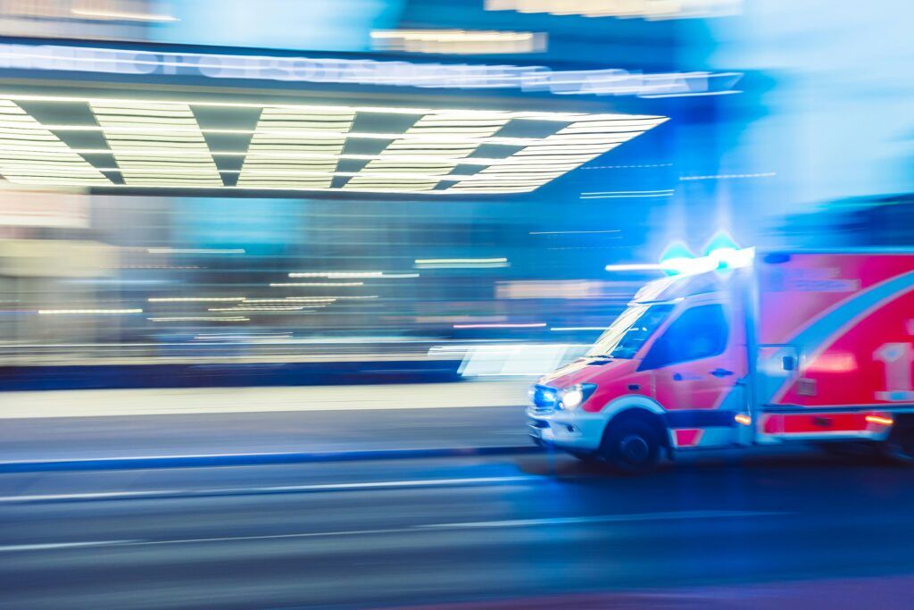 A fast-moving ambulance with flashing blue lights driving through a city street at night, captured with motion blur that emphasizes speed and urgency while bright urban lights streak across the background.