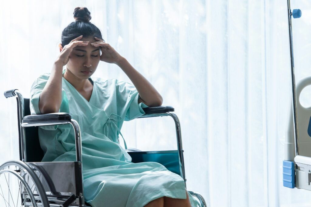 A woman in a hospital gown sitting in a wheelchair, leaning forward with her hands pressed to her forehead, appearing exhausted or in pain as soft daylight filters through sheer curtains behind her.