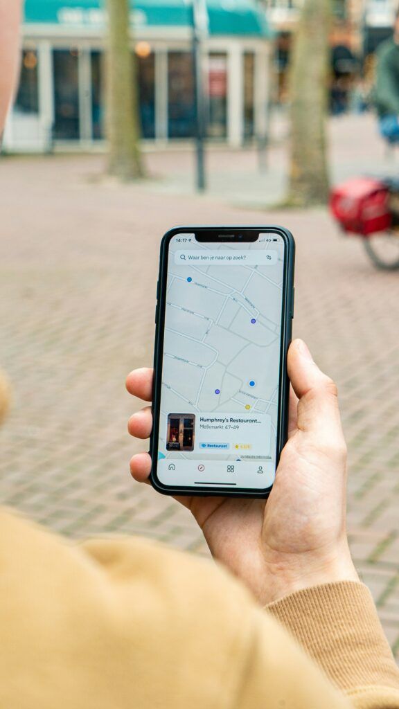A person holding a smartphone in one hand while standing outdoors on a city street, with the screen displaying a map interface showing nearby streets, location pins, and a restaurant listing, while storefronts, trees, and a parked bicycle appear softly blurred in the background.