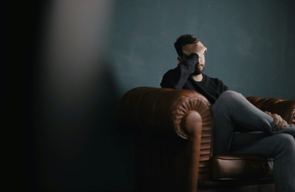 A man sitting alone on a brown leather couch in a dimly lit room, leaning back with one hand covering part of his face as though overwhelmed or deep in thought, with a dark teal wall behind him and soft shadows filling the space.