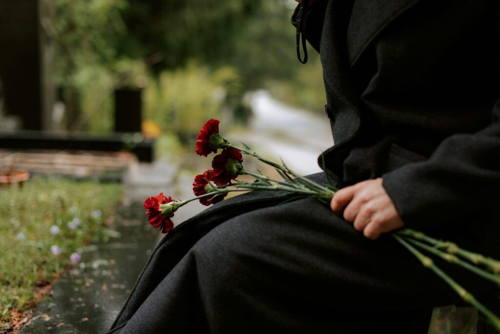 A person dressed in dark clothing sitting in a cemetery, holding a small bundle of red carnations across their lap as the surroundings appear wet and somber, suggesting a moment of mourning.