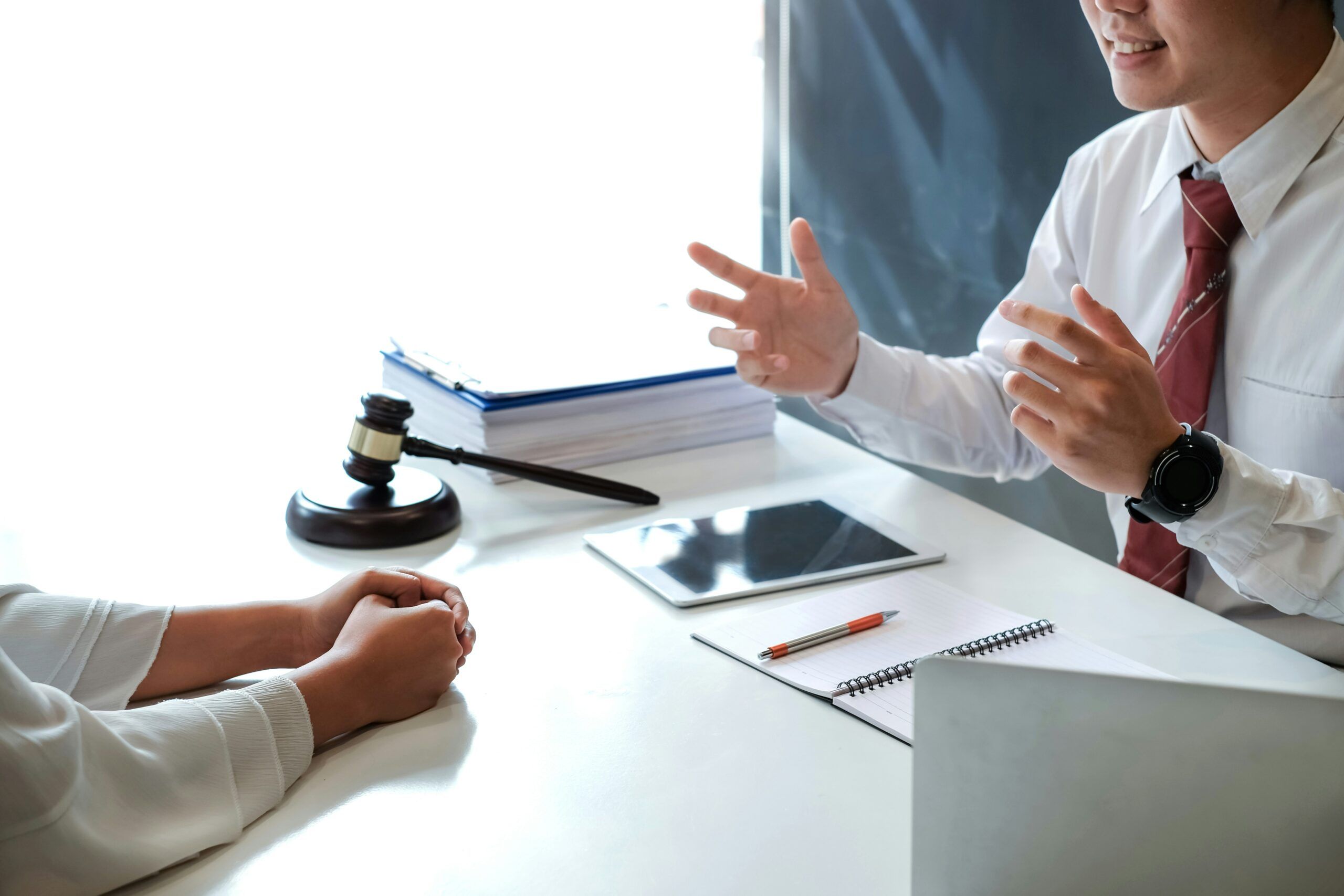 A lawyer gestures while explaining legal matters to a client across a desk, with a gavel, documents, a tablet, and a notebook visible during a professional consultation.