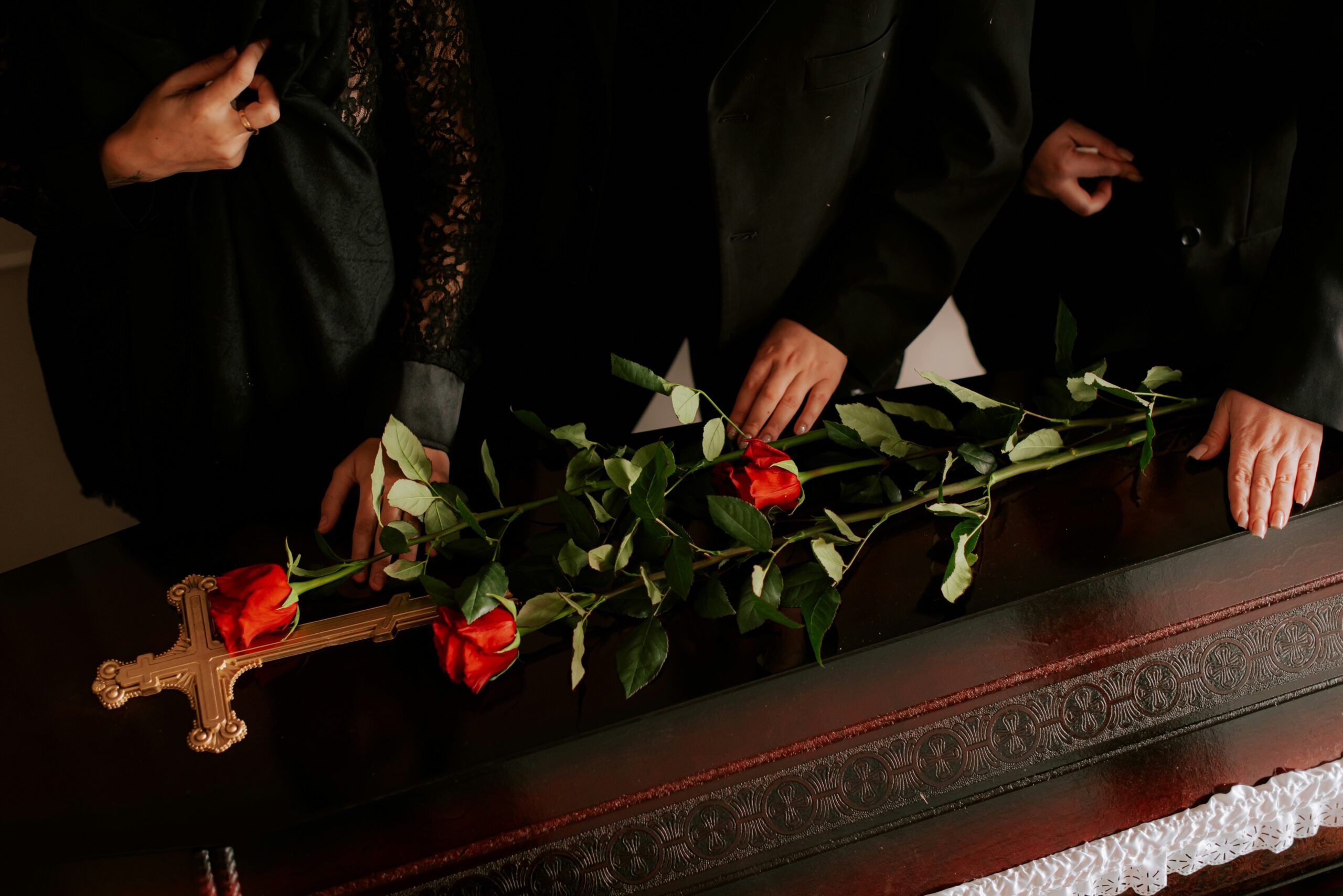 Mourners dressed in black place red roses on top of a coffin during a funeral service, with a gold cross resting near the flowers and hands gently touching the casket in a dimly lit setting.