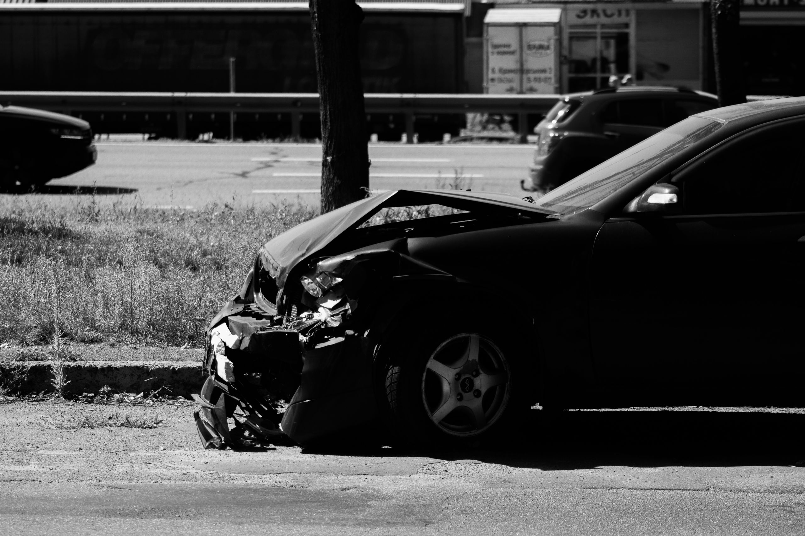 Black-and-white photograph of a car with severe front-end damage parked at the roadside after a collision, showing a crumpled hood, broken bumper, and exposed internal components, with other vehicles and a roadway visible in the background.